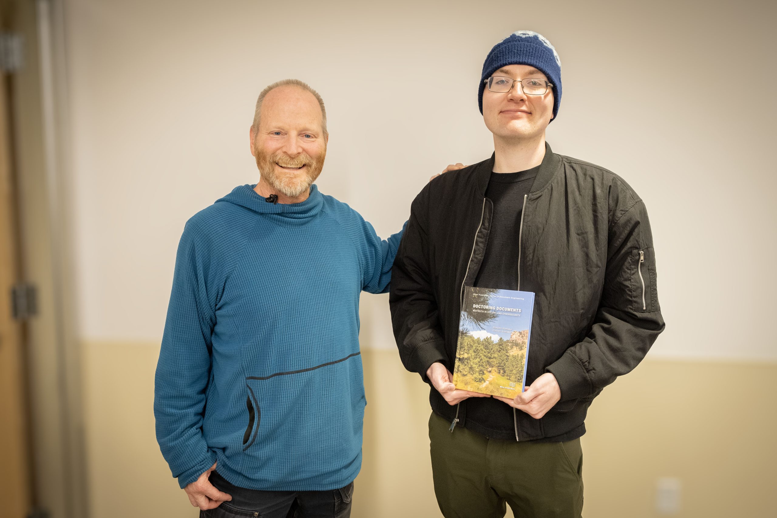 A man in a sweatshirt stands next to another younger man in a beanie hate holding a book.