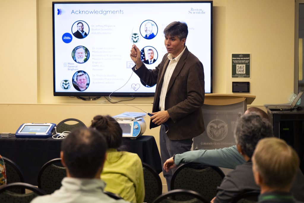 A man in a suit holds up a medical device as he presents about his research to a room of attendees.