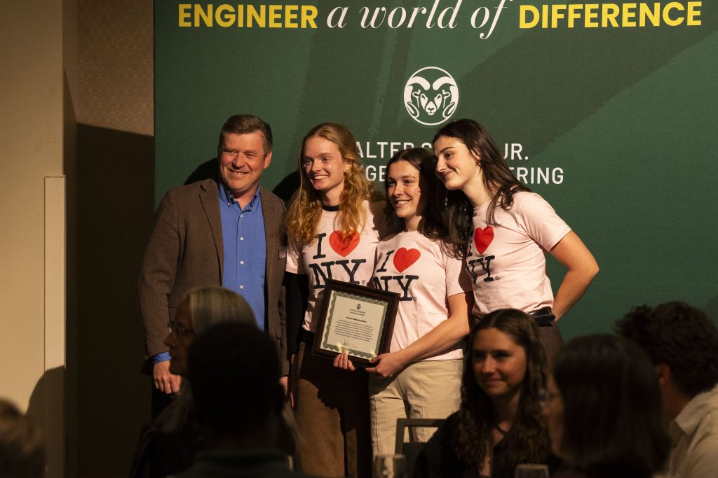 Four people stand for a photo, one man in a suit with three girls who have “I heart N Y” on their shirts. The girls are holding awards. 