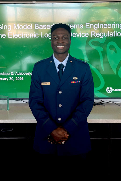 A man in a formal uniform smiles in front of a TV screen.