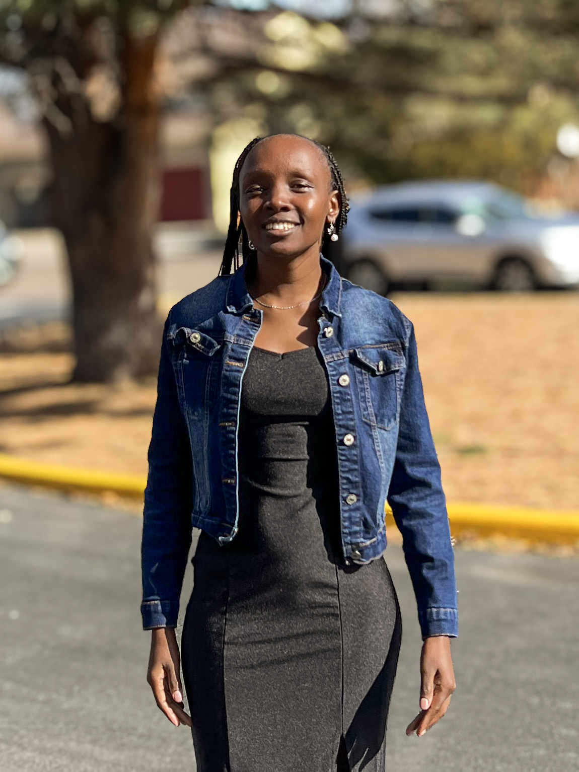 A woman in a dress and jean jacket under a tree in a parking lot.