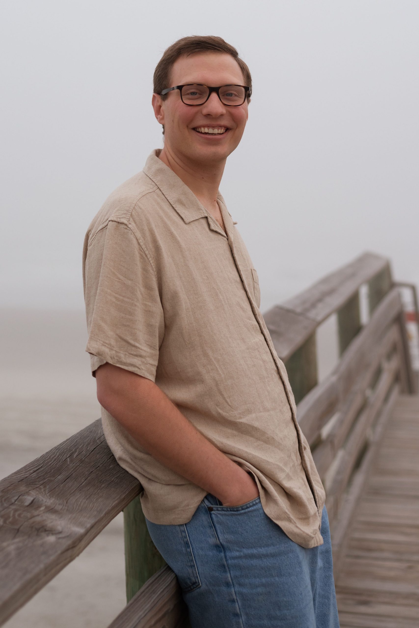A man in jeans and collared shirt leaning on a wooden railing by a beach.