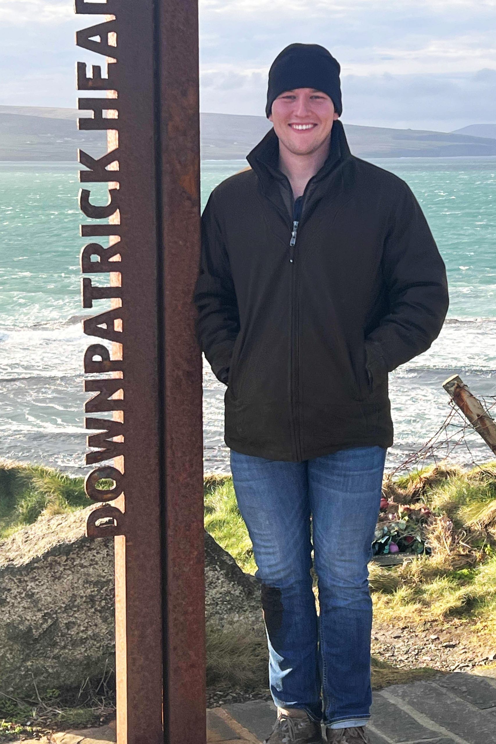 A man in a winter hat, coat, and jeans stands near the ocean next to a metal pole with “Downpatrick head” written on it.