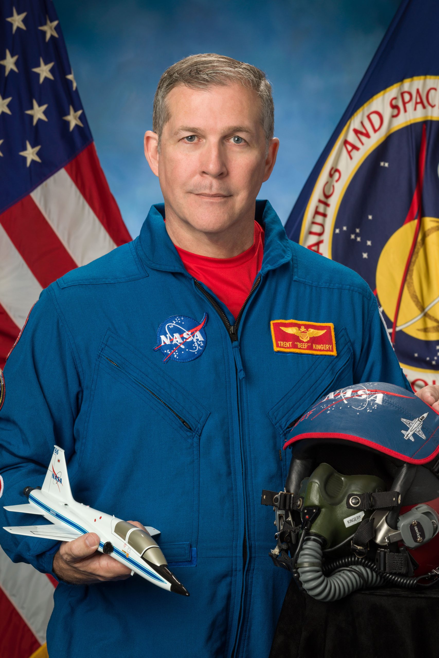 A man in a flight suit holds a model jet plane and a flight helmet.