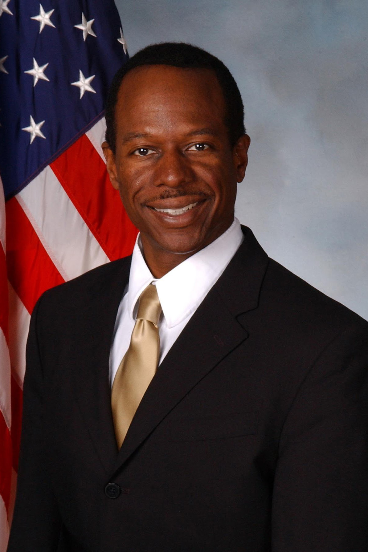A man in a formal business suit with the American flag behind him.