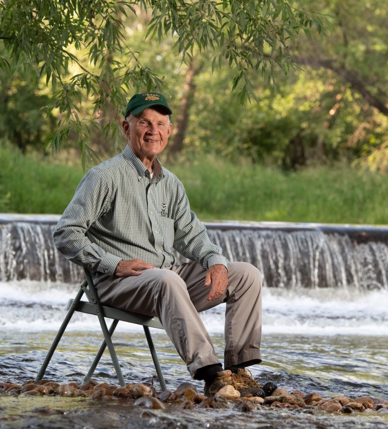 Image of an elderly man in a CSU hat sitting on a folding chair in a river.