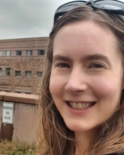 A woman stands outside a building with sunglasses on her head.