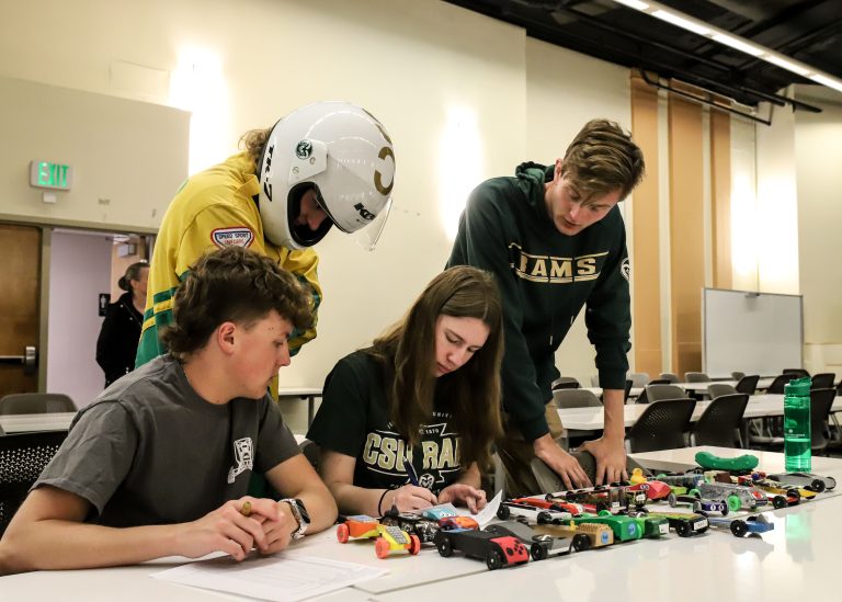 Students standing behind and sitting at a desk work on a project.